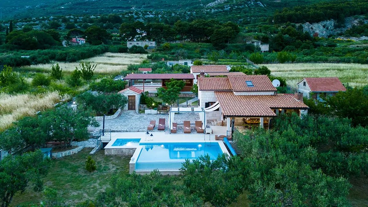 View of the villa at dusk from a semi-bird's eye perspective, showcasing the villa, pool, garden with olive trees.